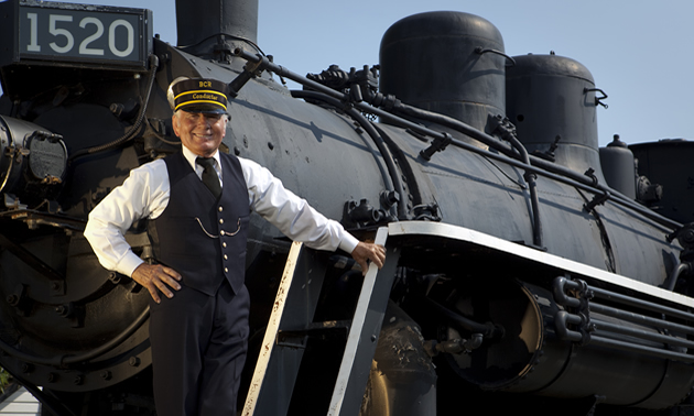 A train conductor stands posed on the front of an antique engine.