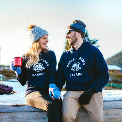 Man and woman sitting together wearing navy blue hoodies with Parks Canada logo. Woman is holding red mug. 