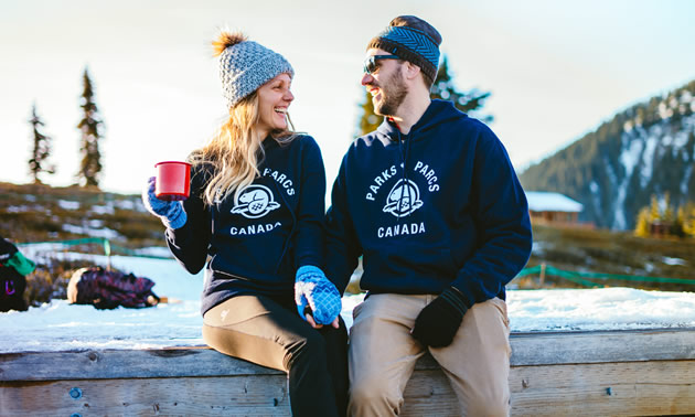 Man and woman sitting together wearing navy blue hoodies with Parks Canada logo. Woman is holding red mug. 