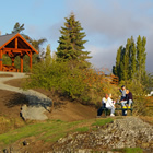 Patrons sit at a picnic table surrounded by green trees, grass, and a far-off gazebo.
