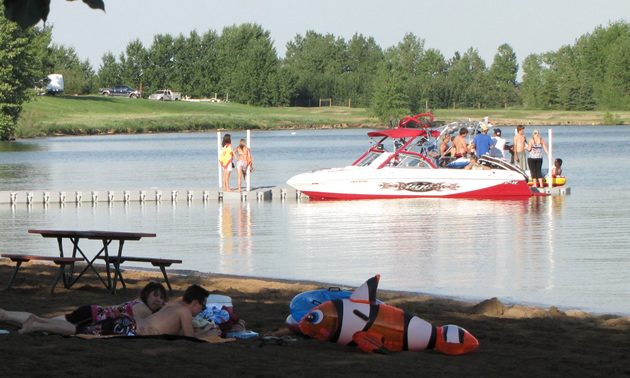 Sunbathers on a beach in the foreground, children and a power boat in the water in the mid-ground and a grassy slope in the far background