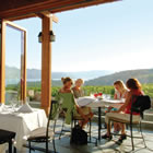 Four women sit at a dining table on a patio overlooking vineyards, lake, low mountains and sky