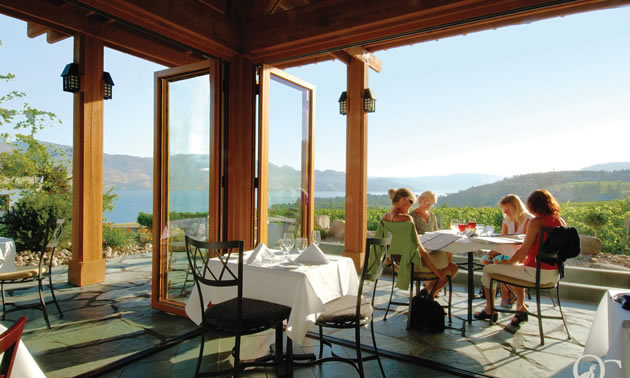 Four women sit at a dining table on a patio overlooking vineyards, lake, low mountains and sky