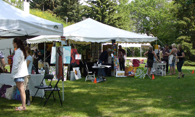 A white tent displays a number of art works and is surrounded by people and green lawn.