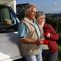 A couple lean against their RV and drink tea against a scenic background.