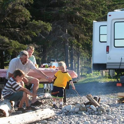 Family sitting at campfire in daytime, with fifth-wheel trailer in background. 