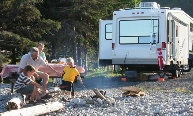 Family sitting at campfire in daytime, with fifth-wheel trailer in background. 