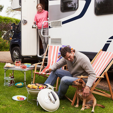Couple In an RV enjoying barbecue on camping holiday.  The man is sitting in a lawn chair leaning over and petting his dog. 