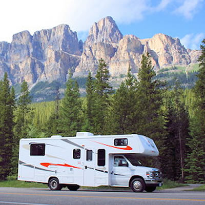 Picture of RV with towering mountains and pine tree in the background. 