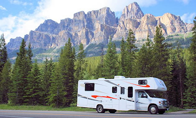 Picture of RV with towering mountains and pine tree in the background.