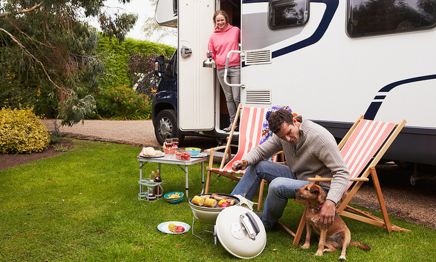Couple In an RV enjoying barbecue on camping holiday. The man is sitting in a lawn chair leaning over and petting his dog.