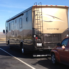 A large brown RV unit pulls a red SUV that displays the Saskatchewan roughriders flag against a background of windmills.