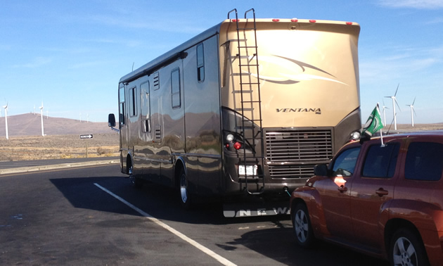 A large brown RV unit pulls a red SUV that displays the Saskatchewan roughriders flag against a background of windmills.