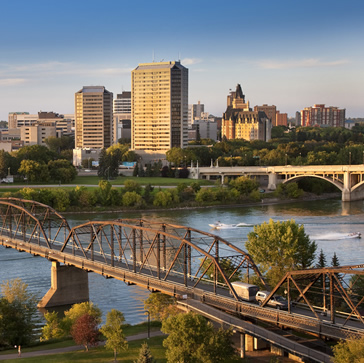 Saskatoon's skyline is illuminated between river and blue sky. A bridge visually leads you into the city.