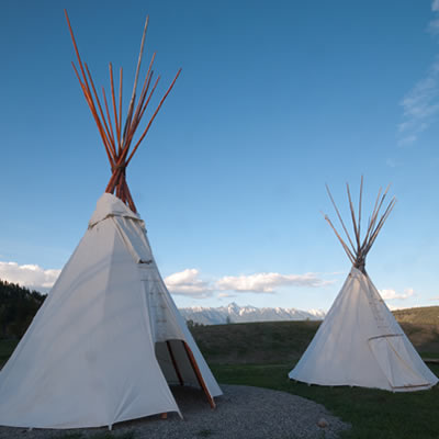 Two teepees made of white canvas, set up in a camp at St. Eugene RV Park, with a view of Rocky Mountains in the distance. 