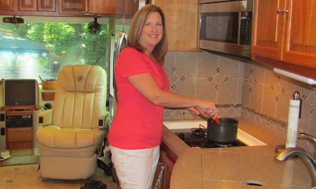 Sylvia stands in her RV kitchen smiling at the camera while cooking in a saucepan.