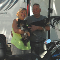 A couple each hold a small dog, standing in front of a dark purple Harley and in front of a new toy-hauler trailer.
