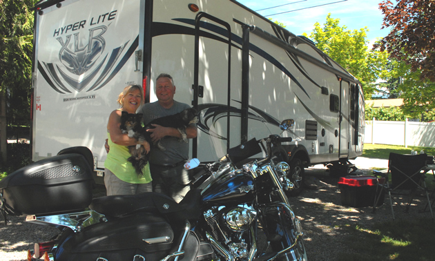 A couple each hold a small dog, standing in front of a dark purple Harley and in front of a new toy-hauler trailer.