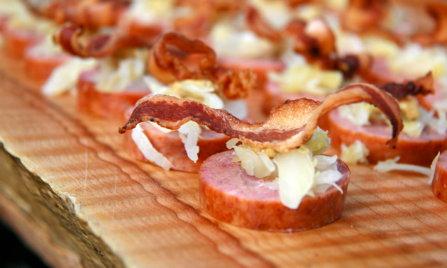 A plank is set with slices of sausage topped with bacon and shavings of cheese.