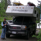 A man stands at the front of a black pickup with an adventure camper uploaded. It has kayaks on top.