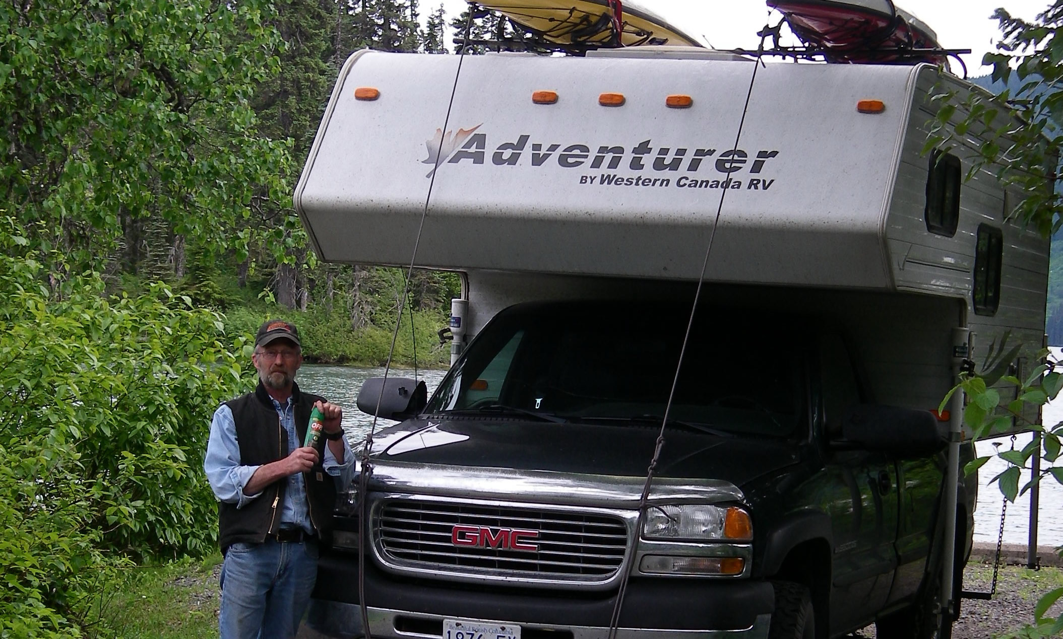 A man stands at the front of a black pickup with an adventure camper uploaded. It has kayaks on top.