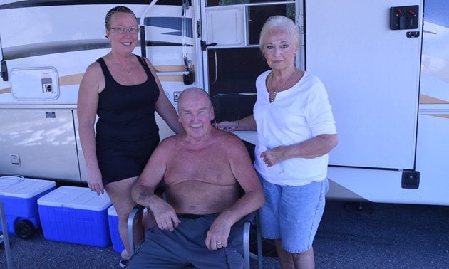 Two women standing, shirtless senior man sitting, with an RV unit behind them