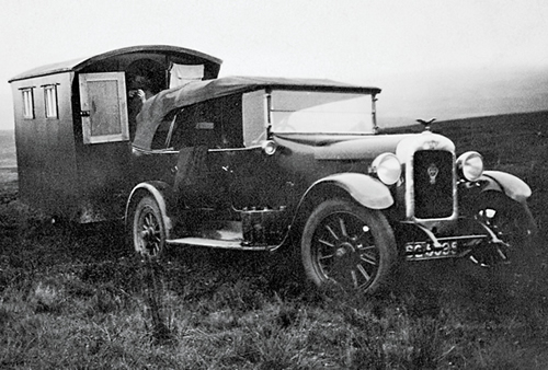 A black and white photo shows a car pulling a wooden travel trailer.