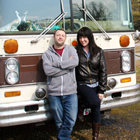 Jeanette wears a leather coat and stands next to Dennis in who wears a hoodie and jeans. Behind them is the brown and cream bumper of their flat-nose bus.