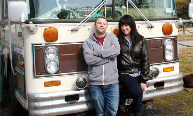 Jeanette wears a leather coat and stands next to Dennis in who wears a hoodie and jeans. Behind them is the brown and cream bumper of their flat-nose bus.