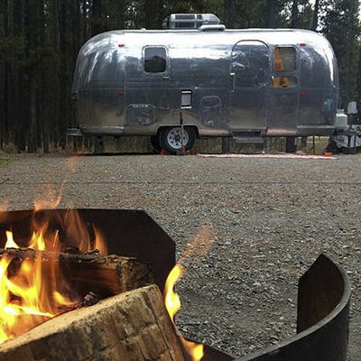 Picture of campfire in foreground, with silver Airstream trailer in background. 