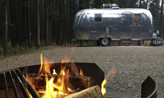 Picture of campfire in foreground, with silver Airstream trailer in background. 