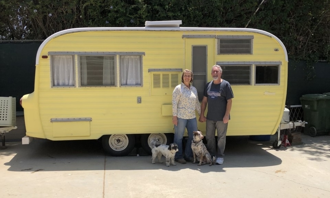 Couple with two dogs standing in front of a yellow vintage RV