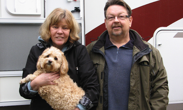 middle-aged couple standing in front of RV; woman holding a dog.