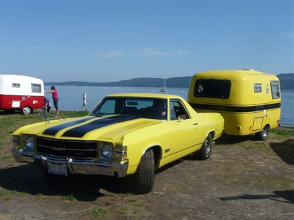 A restored black and yellow Boler trailer, towed by a black and yellow El Camino sits on display at a beachside parking lot.