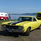 A restored black and yellow Boler trailer, towed by a black and yellow El Camino sits on display at a beachside parking lot.