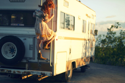 A young woman in a dress climbs down a ladder that is attached to the end of an RV.