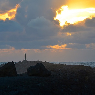 A lighthouse on the edge of a rock jutting out into the coast near Rockaway Beach, Oregon.  There is a black stormy sky, with one opening in the clouds showing light. 