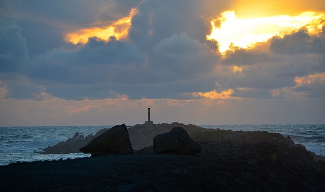 A lighthouse on the edge of a rock jutting out into the coast near Rockaway Beach, Oregon. There is a black stormy sky, with one opening in the clouds showing light.