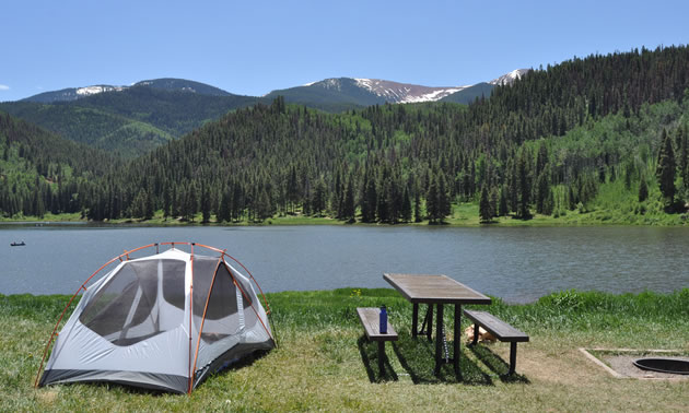 Scenic picture of tent, picnic table, with lake and mountains in background. 