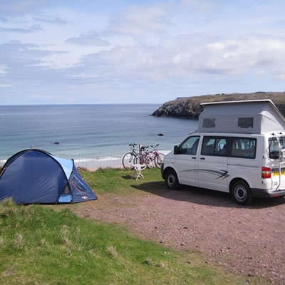 Blue tent and white pop-up van parked along cliff overlooking ocean, bikes parked along a fence.  