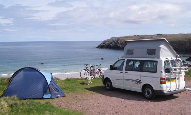 Blue tent and white pop-up van parked along cliff overlooking ocean, bikes parked along a fence.  