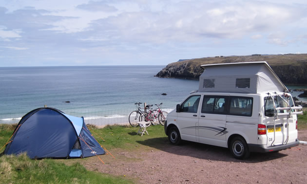 Blue tent and white pop-up van parked along cliff overlooking ocean, bikes parked along a fence.