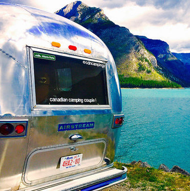 The back end of Dave Barton and Anne Bovan's vintage airstream.  Just beyond their trailer is a green coloured lake and mountains in the background. 