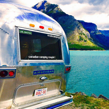 The back end of Dave Barton and Anne Bovan's vintage airstream. Just beyond their trailer is a green coloured lake and mountains in the background.