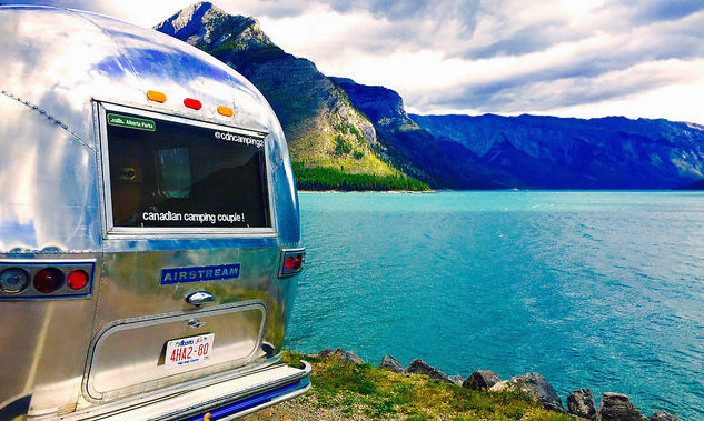 The back end of Dave Barton and Anne Bovan's vintage airstream.  Just beyond their trailer is a green coloured lake and mountains in the background. 