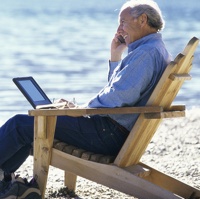 An elderly man sitting in a wooden beach chair talking on his cell phone.  