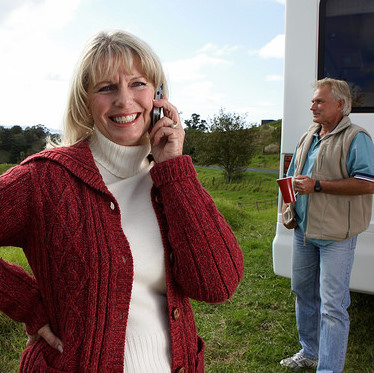 A smiling women pictured in the forground is talking on her cell phone while her husband, who is seen in the backround, is holding a cup of coffee next to their RV.