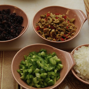 The fresh ingredients of this recipe shown chopped up in small bowls, green pepper, olives, raisins, onions and tomato sauce.