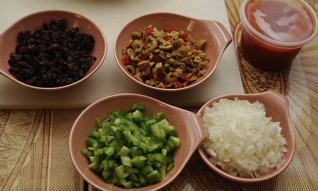 The fresh ingredients of this recipe shown chopped up in small bowls, green pepper, olives, raisins, onions and tomato sauce.