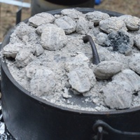 Two dutch ovens on a grate.  One is open and shows a finished pot of potatoes, onions and cheese.  The other pot is closed and has briquettes on top. 